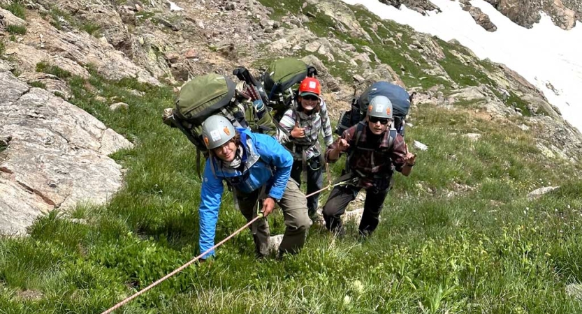 three people wearing safety gear hold onto a rope as the ascend a grassy incline